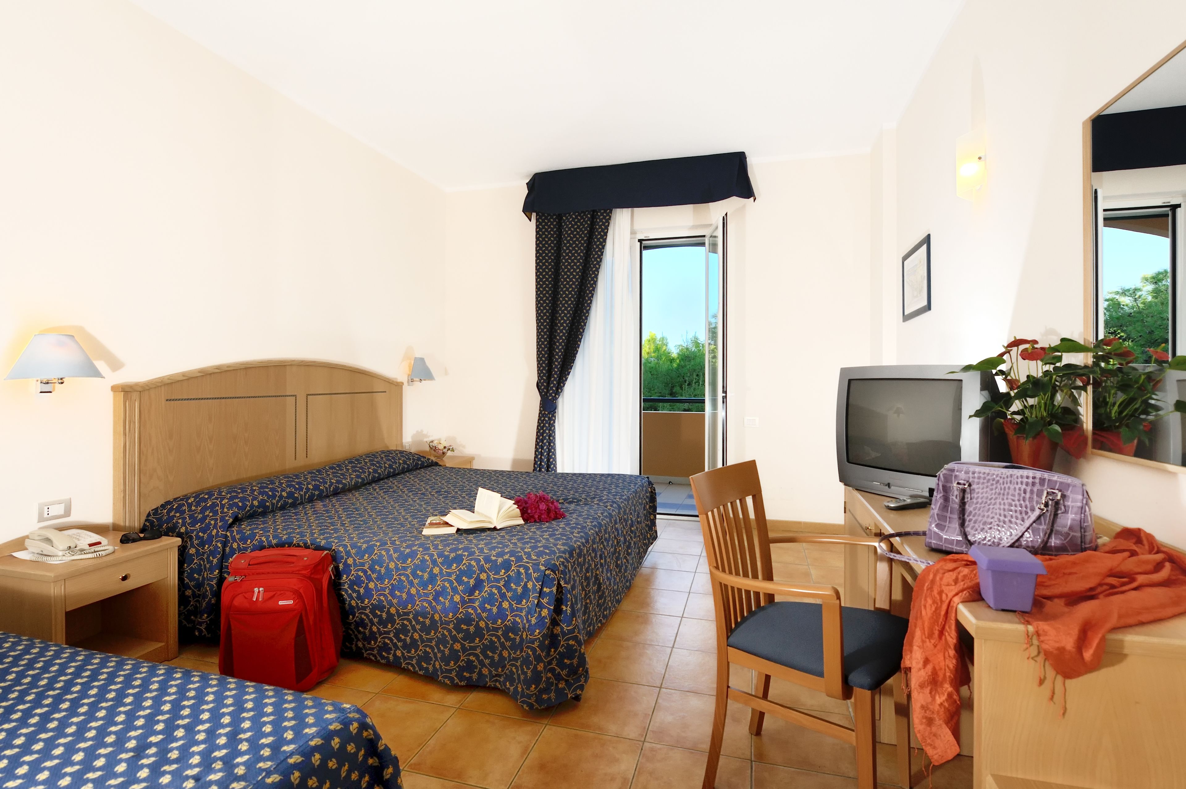 A hotel room with two beds, a desk with a television, a chair, a suitcase, a lamp, and a window with curtains.