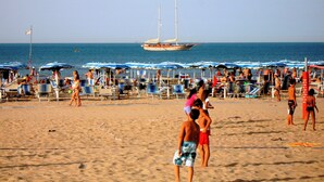 Una playa con hileras de sombrillas y sillas, un velero a lo lejos, y gente disfrutando.