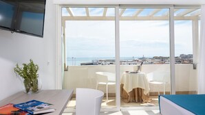 Un balcón con mesa y sillas, un televisor instalado en la pared, y vistas al mar y al paisaje urbano.