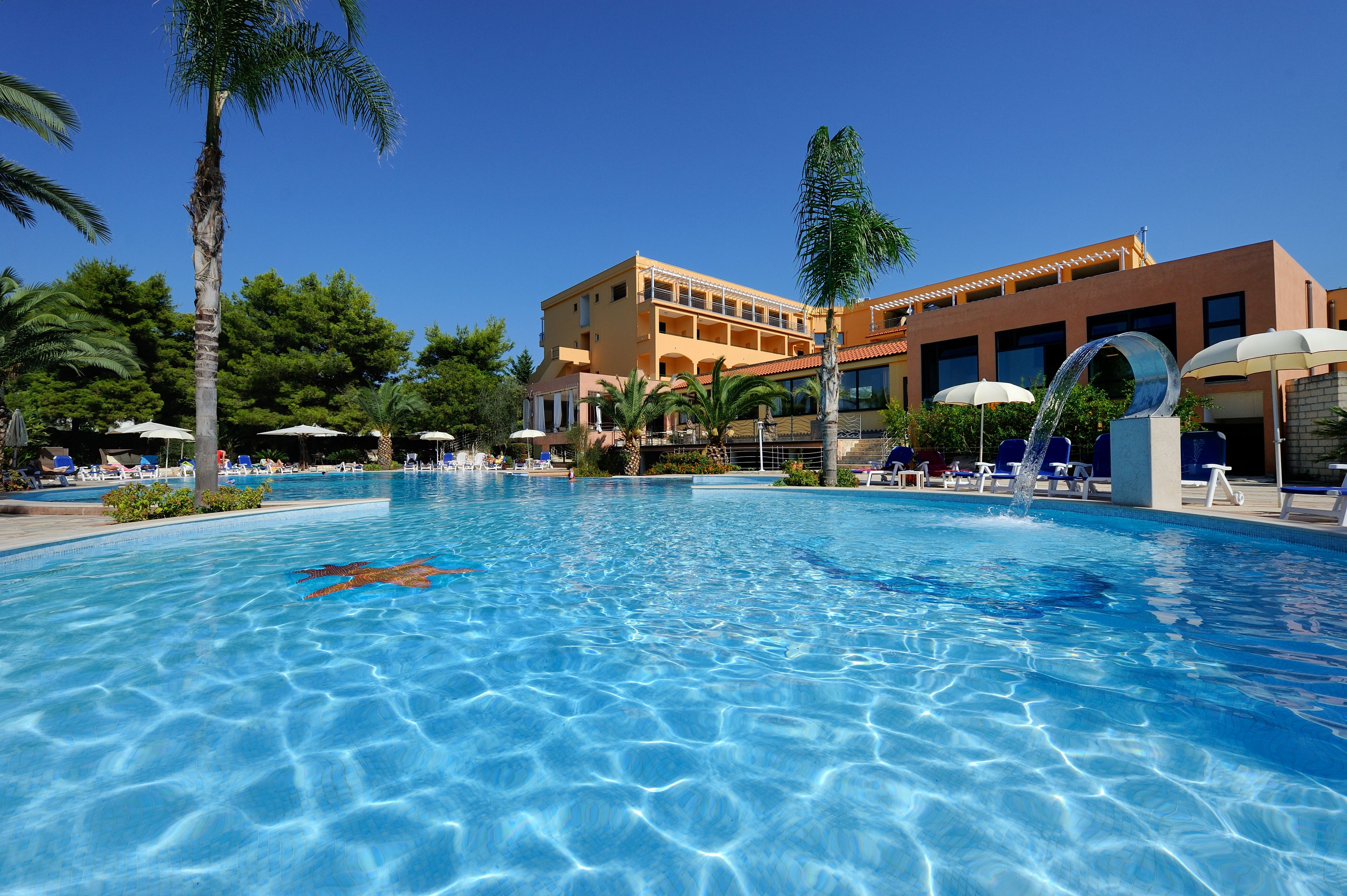 Piscina azul cristalina con sillas de descanso y sombrillas, rodeada de palmeras y un edificio con varios balcones.