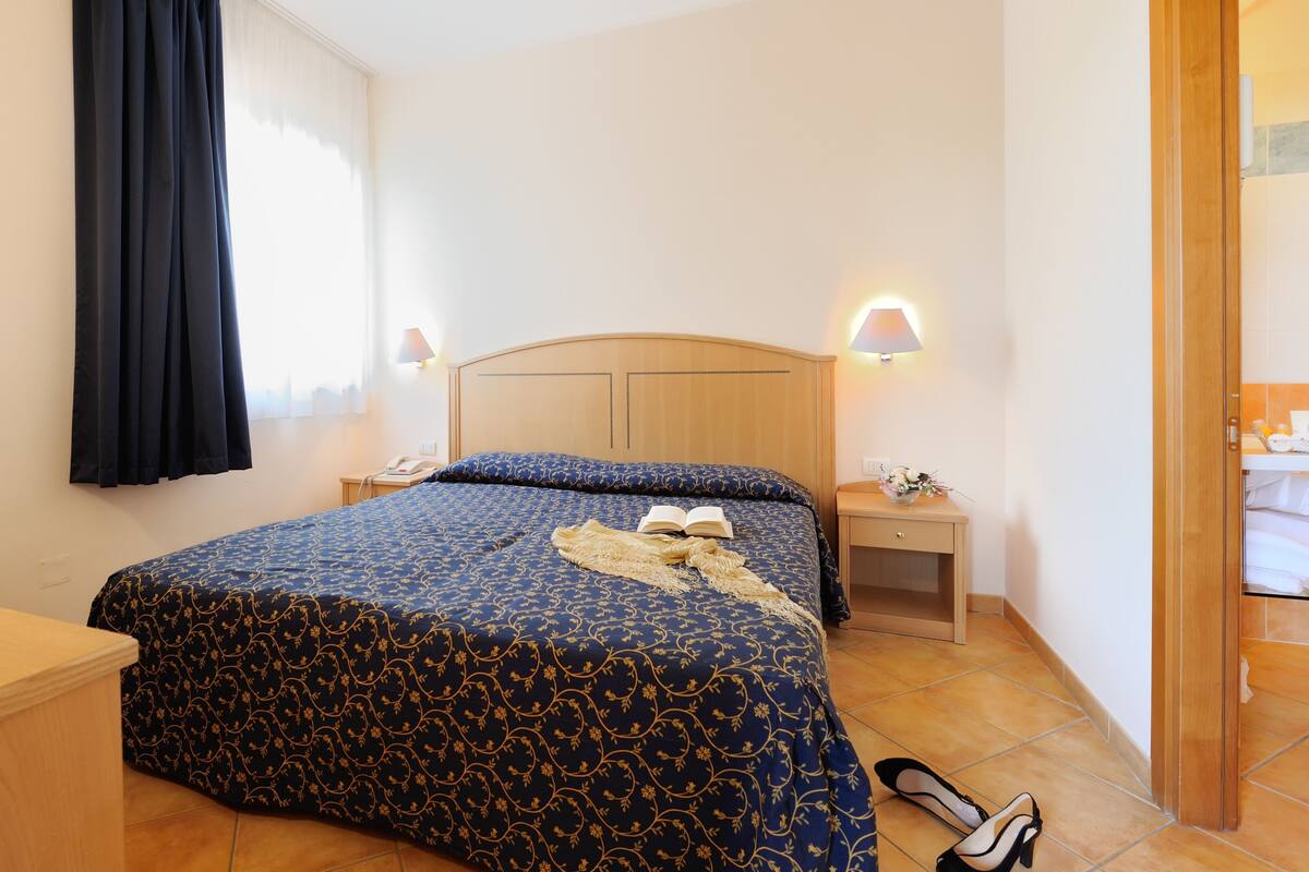 A neatly made bed with a patterned bedspread, a bedside lamp, and a pair of shoes on the floor.