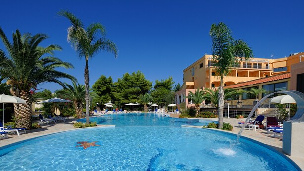 Una piscina con sillas de descanso y sombrillas, rodeada de palmeras y un edificio con balcones.