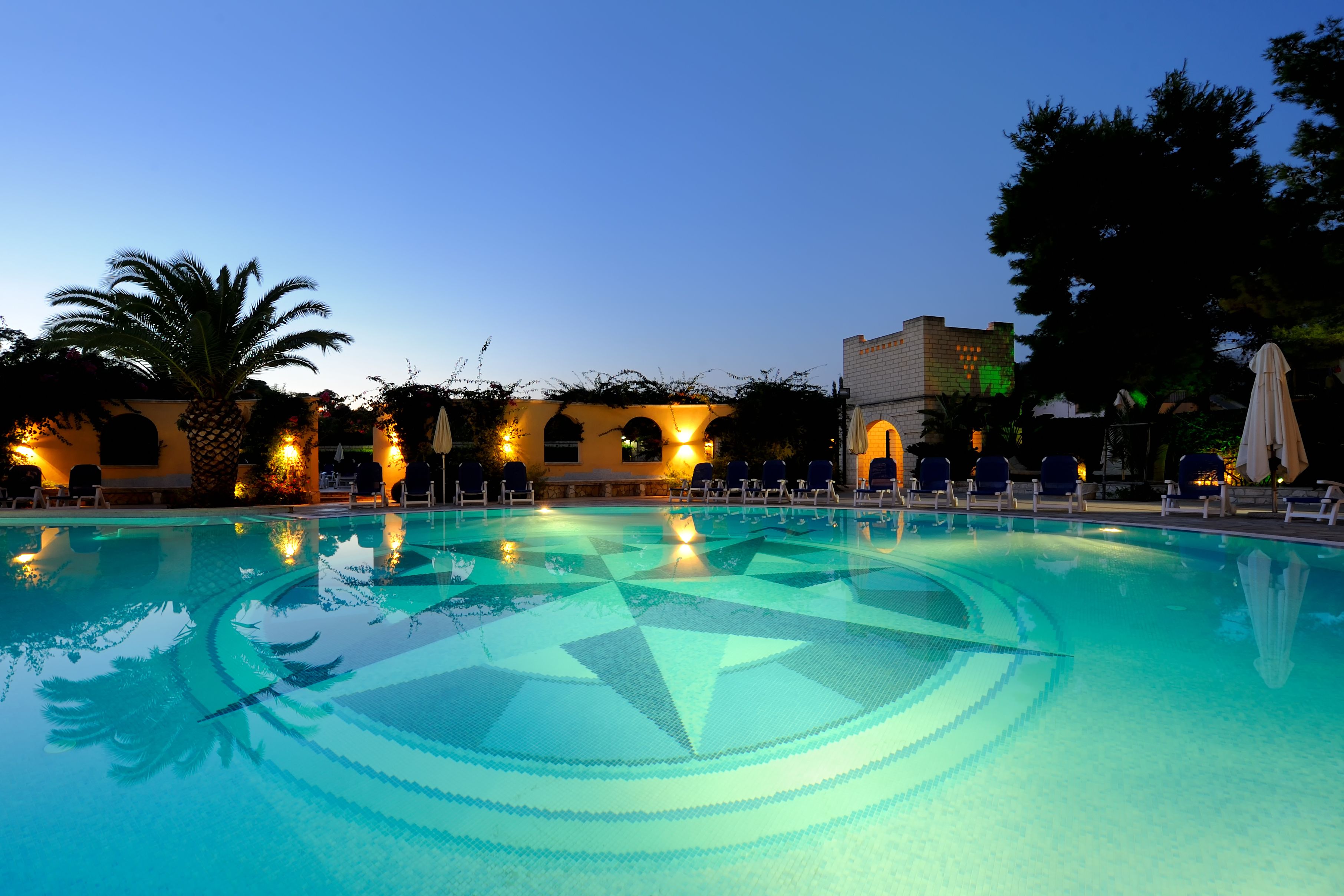 Una piscina con un diseño geométrico, rodeada de sillas de descanso y palmeras, bajo un cielo despejado.
