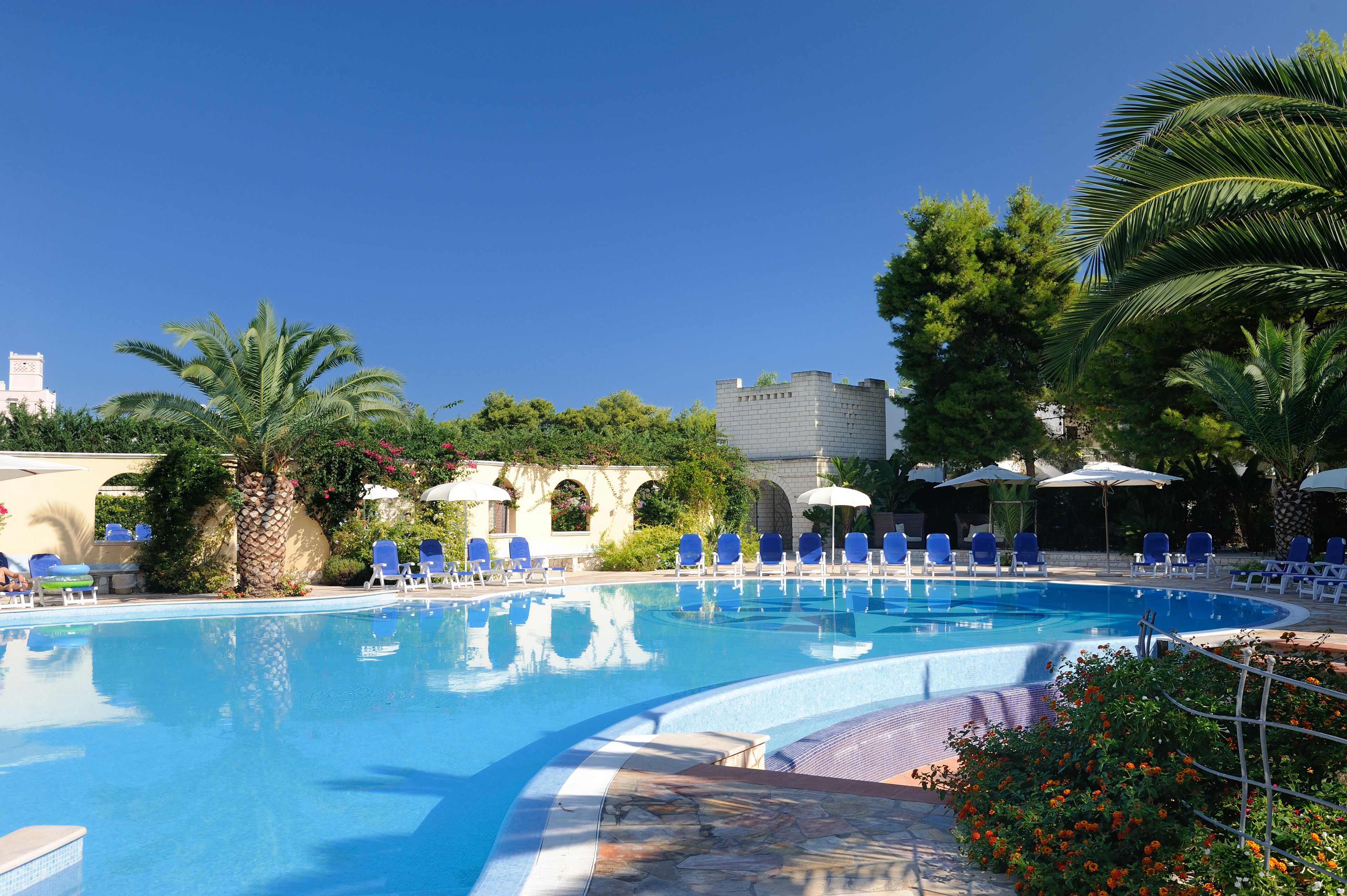 Ein Swimmingpool mit blauem Wasser, umgeben von Liegestühlen und Sonnenschirmen, mit Palmen und einem schlossartigen Gebäude im Hintergrund.