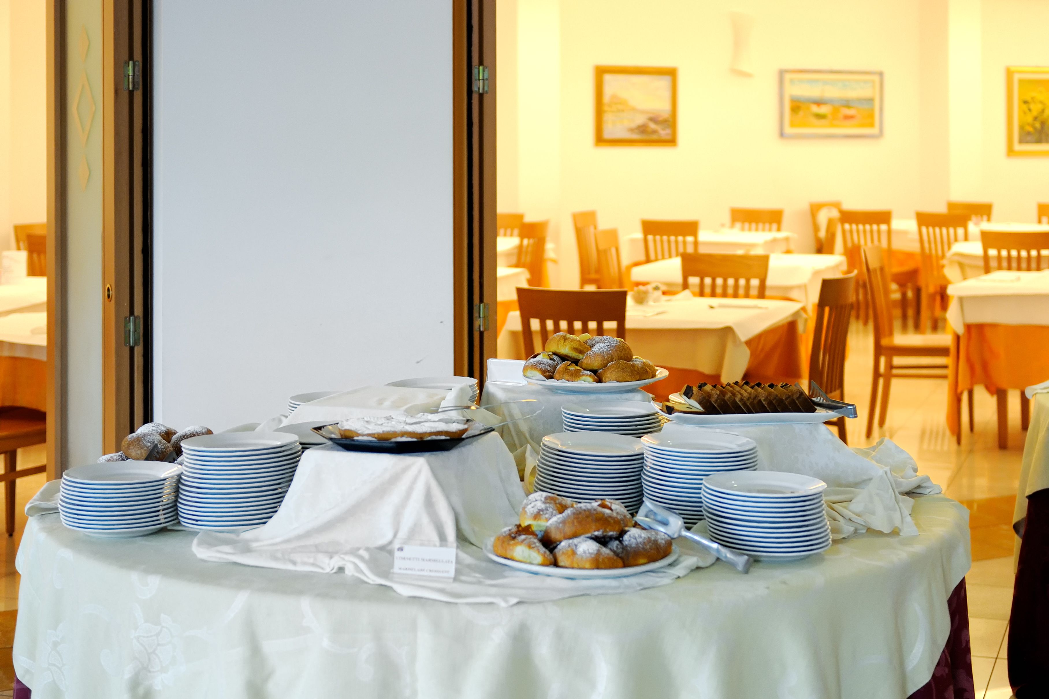 A buffet table with various pastries and stacked plates, set in a dining area with chairs and tables.
