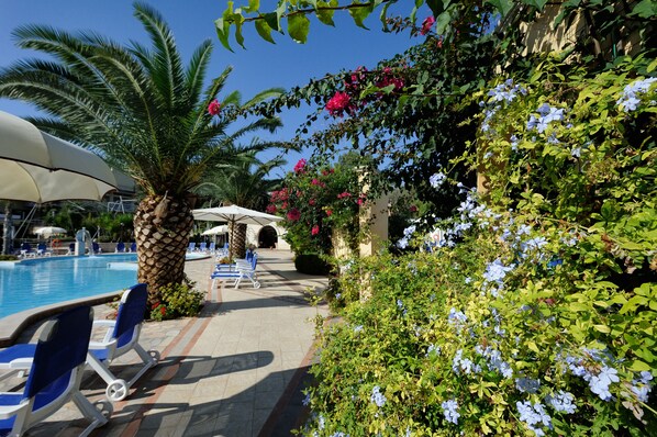 Un área junto a la piscina con sillones de descanso, una palmera y plantas con flores.