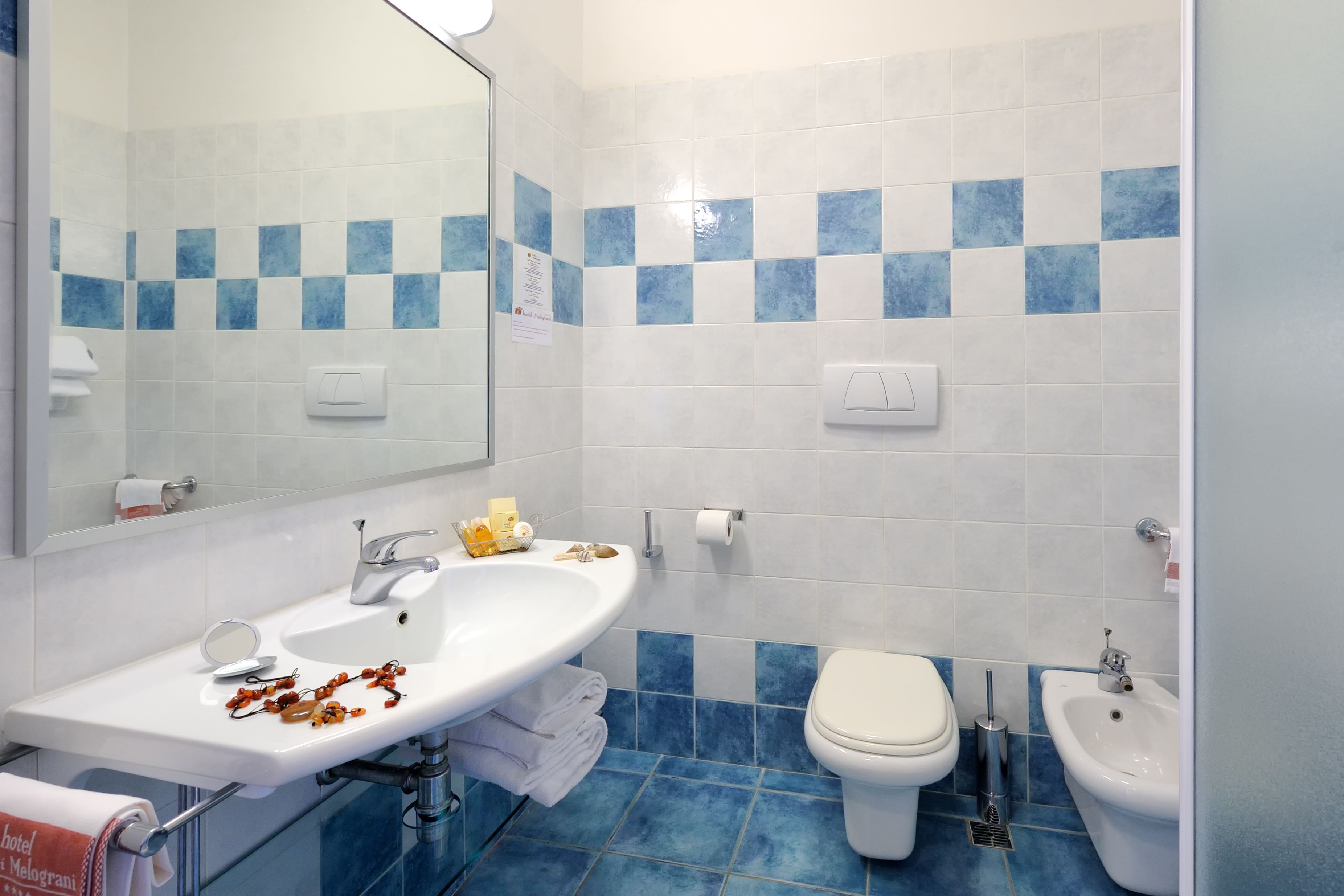 A bathroom with a white sink, mirror, and two sinks; a toilet and bidet; blue and white tiled walls; and a towel rack.
