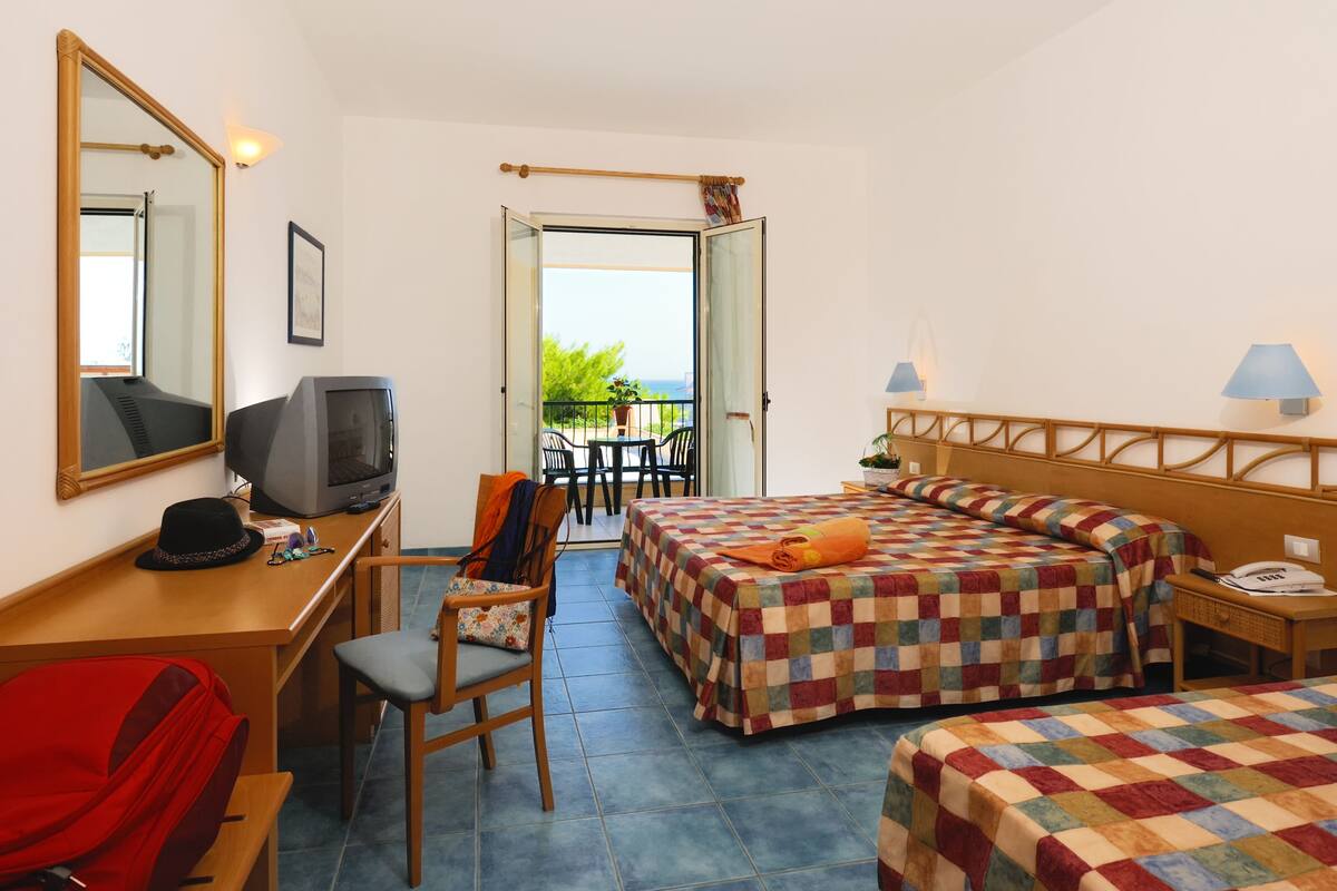 A hotel room with a bed, a desk with a television, a chair, a red suitcase, and a balcony with a view.