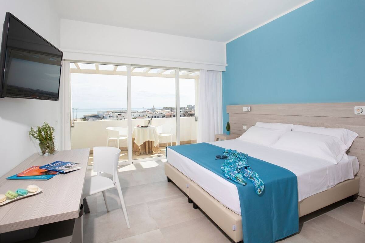 A hotel room with a bed, a desk with a chair, a TV mounted on the wall, and a balcony with a view of the city.