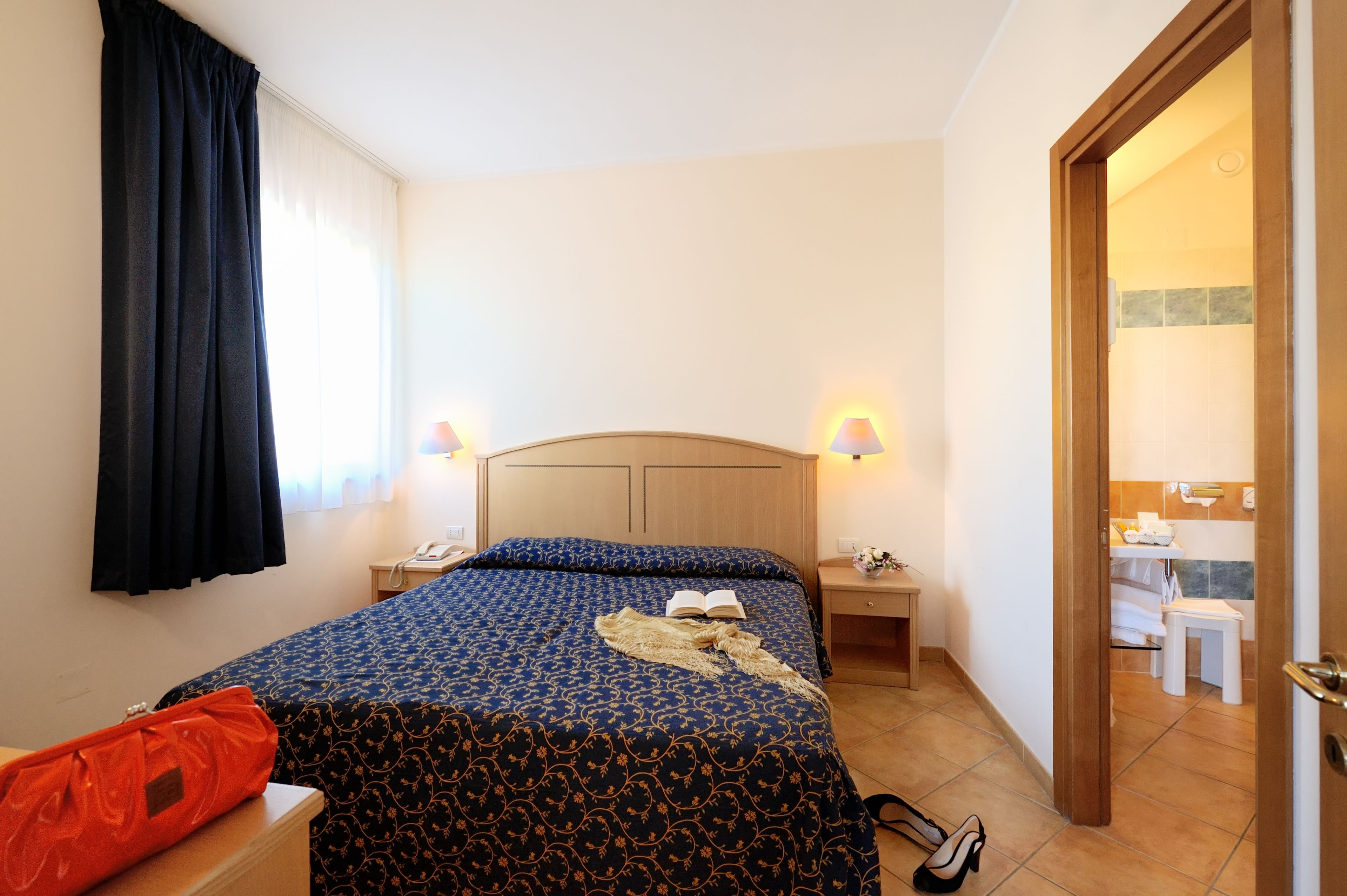 A hotel room with a bed, bedside tables, a nightstand, a chair, and a bathroom visible through an open door.