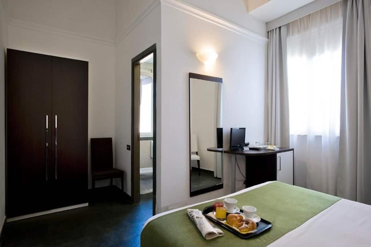 Habitació d'hotel amb llit, escriptori, cadira, armari i un bany visible a través d'una porta oberta.