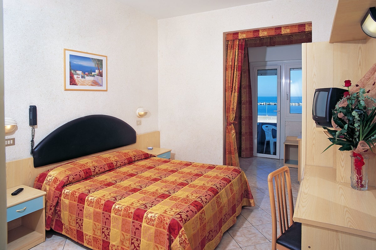 A hotel room with a bed, a desk, a chair, a TV, and a view of the sea through a door.