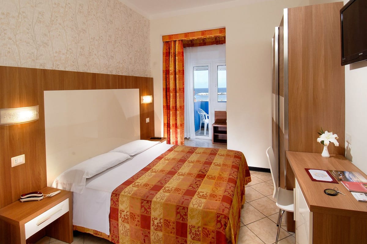 A hotel room with a bed, a desk, a TV, and a view of the sea through a door.