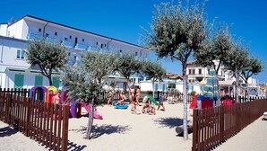 Zona de playa con valla de madera, estructuras de juego coloridas y una hilera de edificios blancos con balcones.