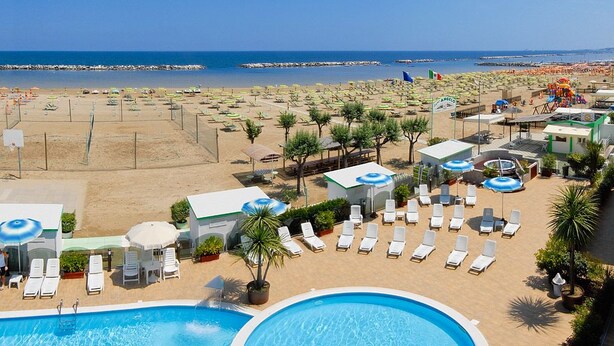 Zona de piscina con sillas de descanso y sombrillas, una playa al fondo y cielo despejado.