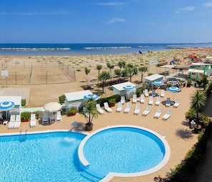 Zona de piscina con sillas de descanso y sombrillas, una playa al fondo y cielo despejado.
