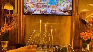 A bar area with a television displaying a scenic view, a bottle of white wine, and two hands holding wine glasses. - Ayasofya Hotel - Special Class (Istanbul)