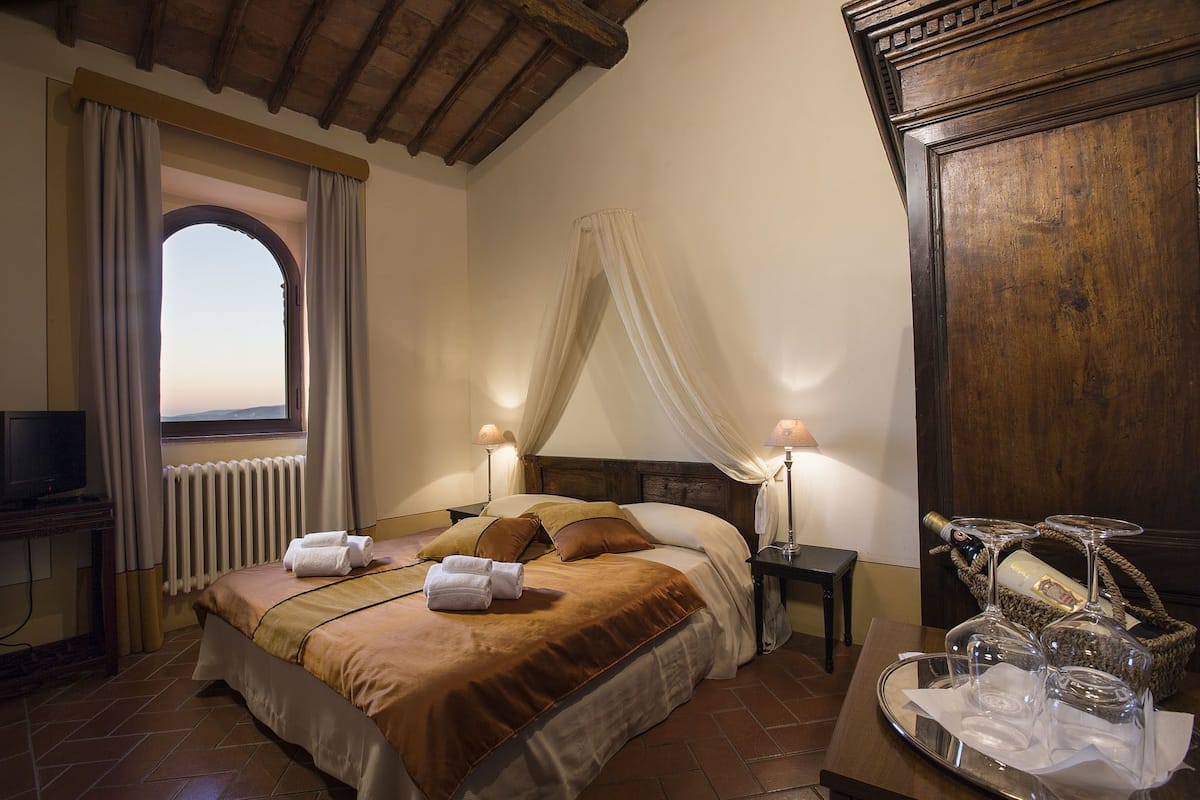 Una camera d'albergo con un letto, due lampade ai lati, una televisione, una finestra con tende, un armadio in legno e un vassoio con bicchieri e una bottiglia.