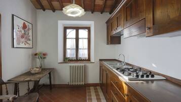 A kitchen with wooden cabinets, a stove, a window with lace curtains, a radiator, and a rustic wooden table with flowers.