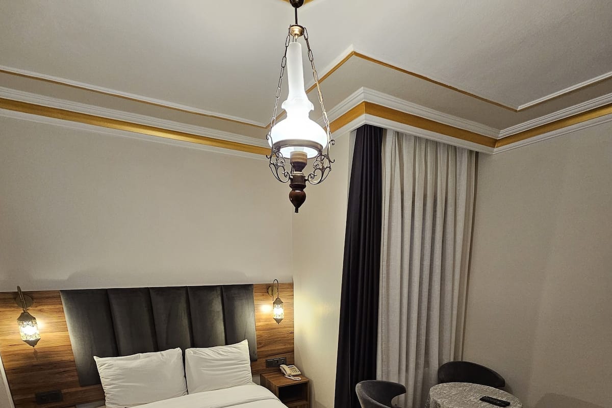 Une chambre d’hôtel avec un grand lit, un parquet en bois, un lustre, une petite table, une chaise et une table de chevet avec un téléphone.