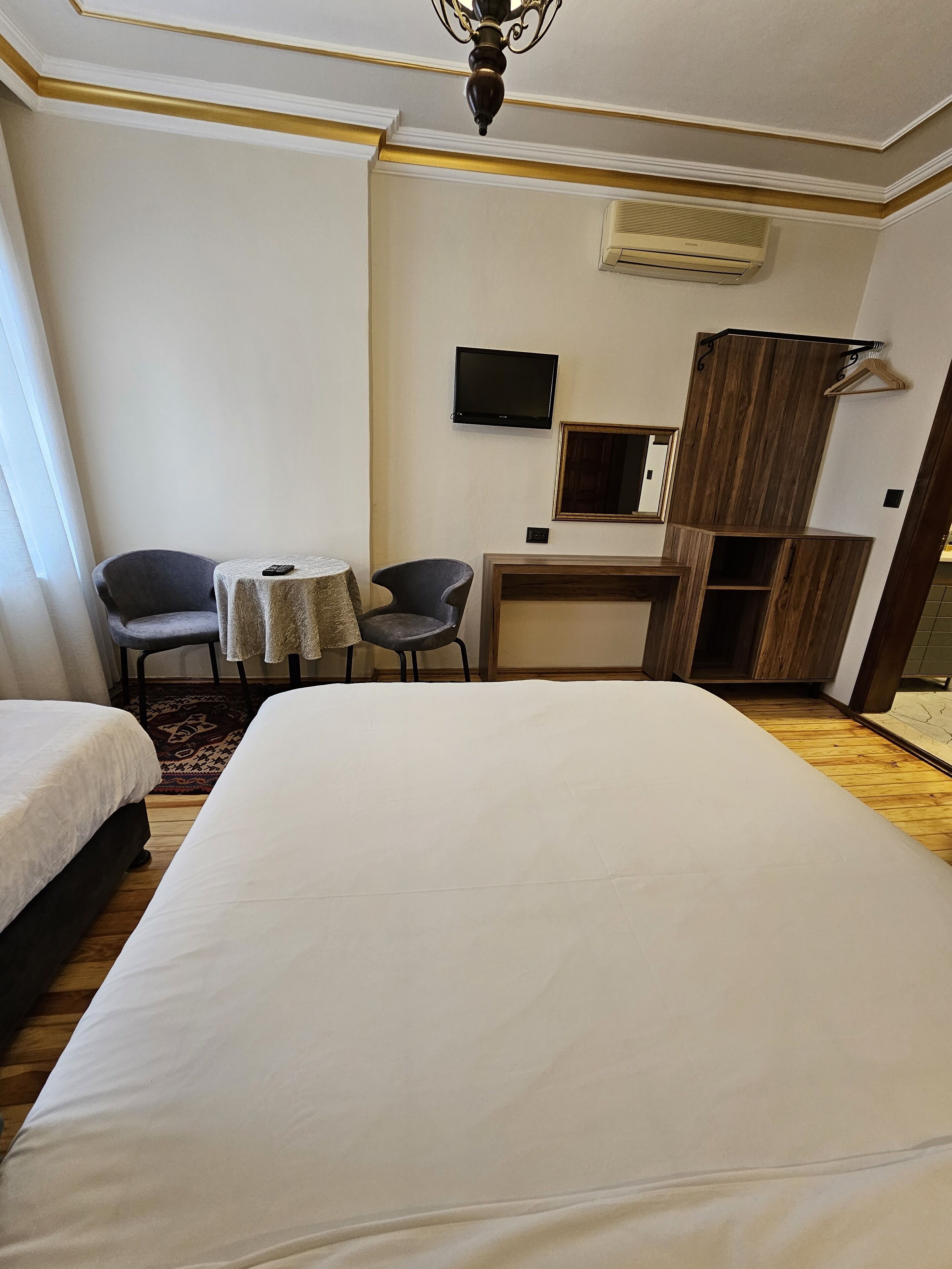 A hotel room with a large bed, two armchairs, a small table, a TV, an air conditioning unit, and a wooden cabinet.