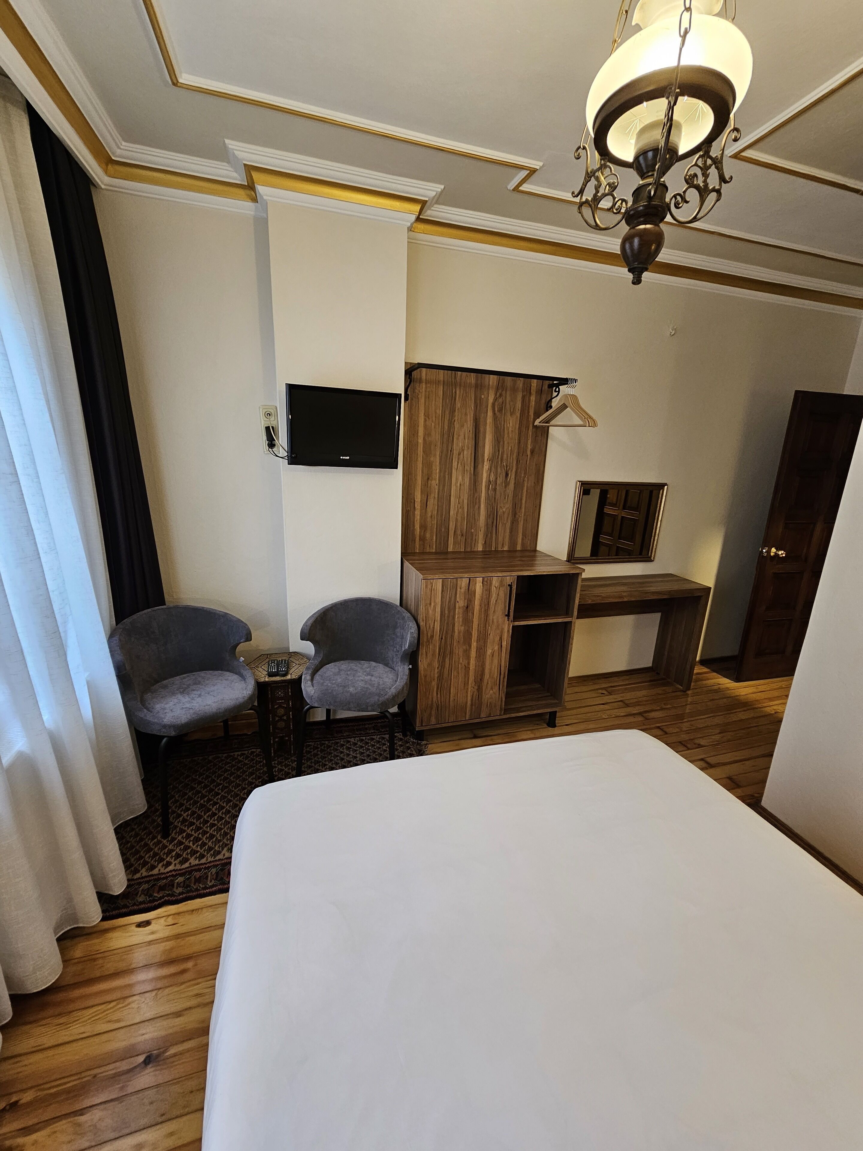 A hotel room with a bed, two armchairs, a wooden wardrobe, a desk, a television, and a chandelier.