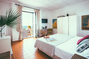 A hotel room with a bed, a desk, a chair, a potted plant, a television, and a view of the sea through the window. - Hotel Sirenetta (Rome)