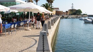 Ein Essbereich am Wasserufer mit Außenbestuhlung und Sonnenschirmen, einer Steinmauer entlang des Wassers und in der Nähe liegenden Booten.