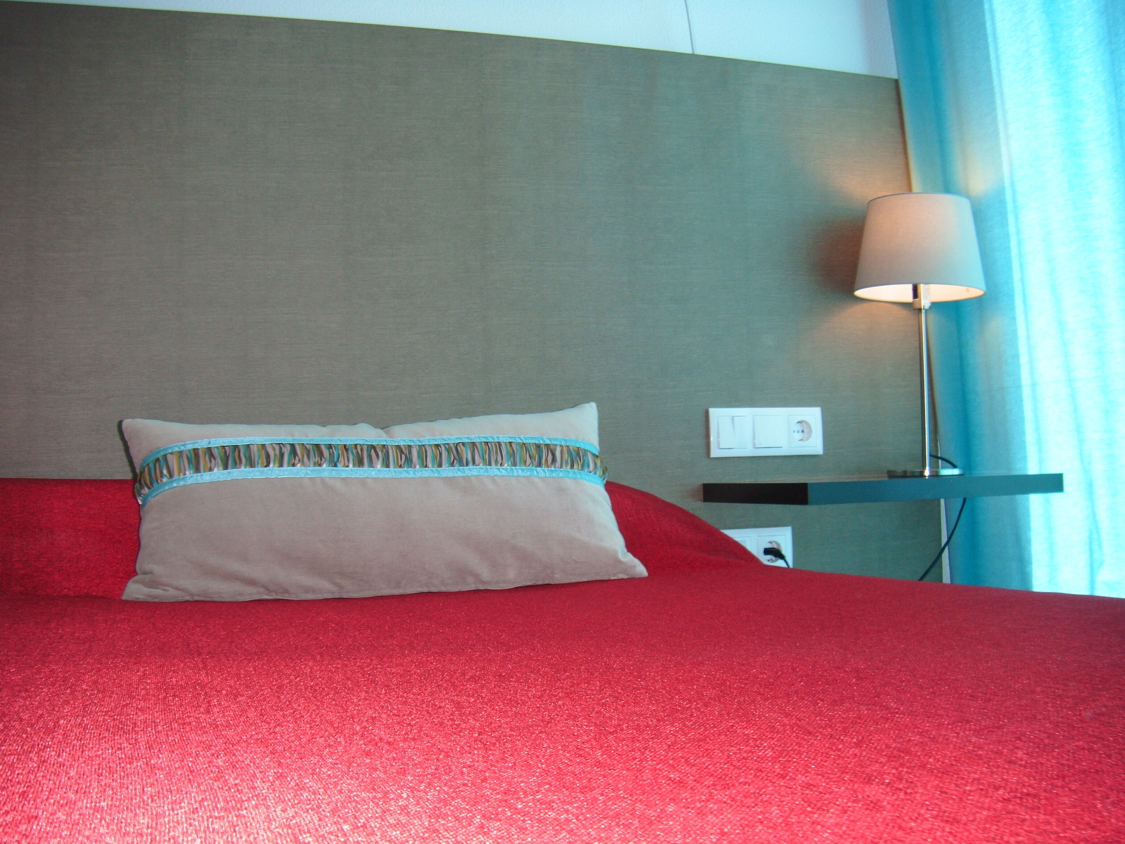 Una cama con cubre cama rojo, un almohadón con detalle estampado en el borde, una lámpara de noche y un enchufe de pared.