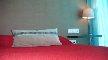 Una cama con cubre cama rojo, un almohadón con detalle estampado en el borde, una lámpara de noche y un enchufe de pared.