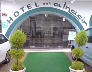 A hotel entrance with a sign reading "HOTEL alnacir" and a 24-hour service symbol. There are two potted trees in front of the entrance. - Alnacir (Faro)