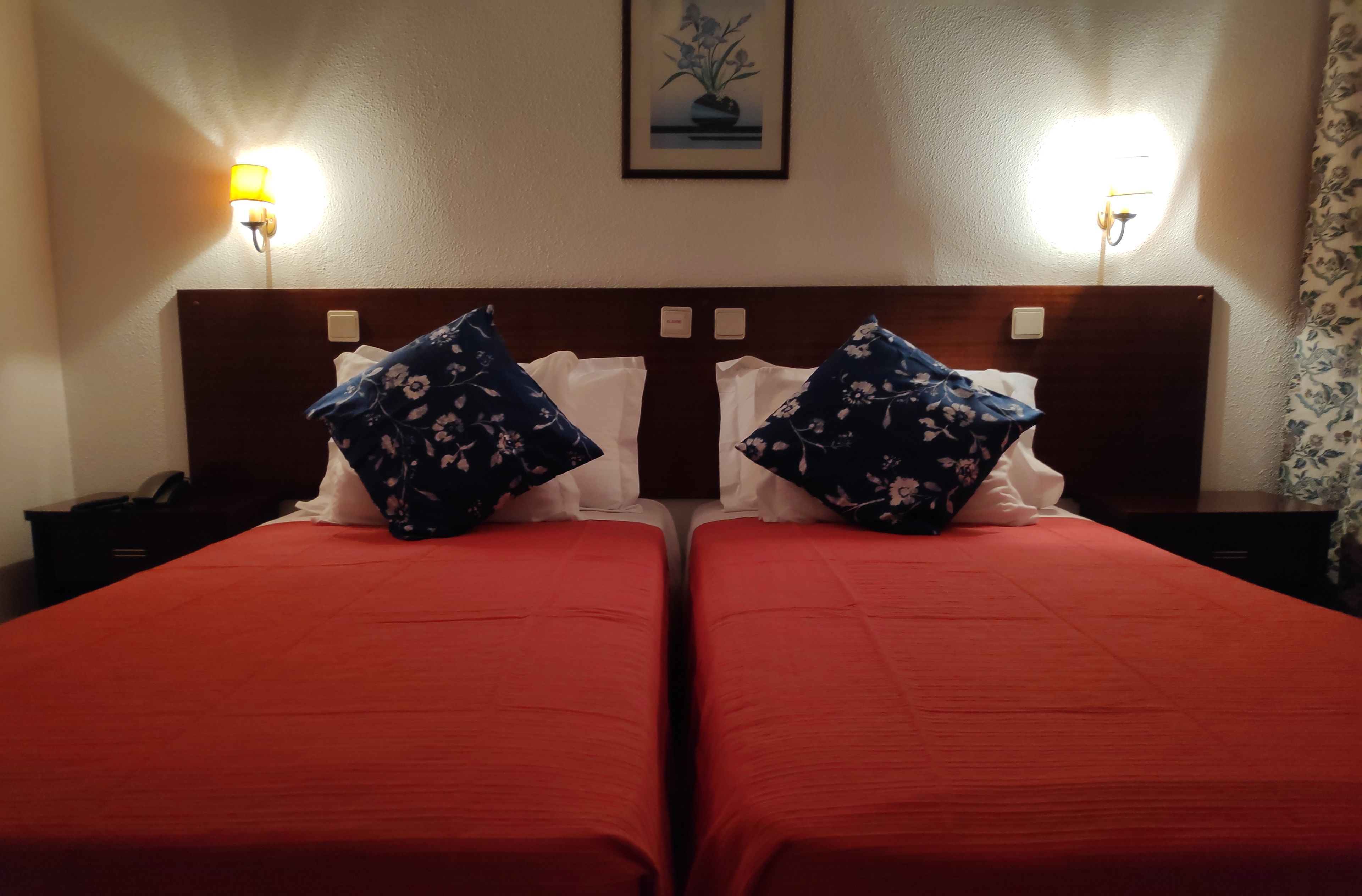 A hotel room with two beds, each with red bedding and floral-patterned pillows. There are bedside lamps and a framed picture on the wall.