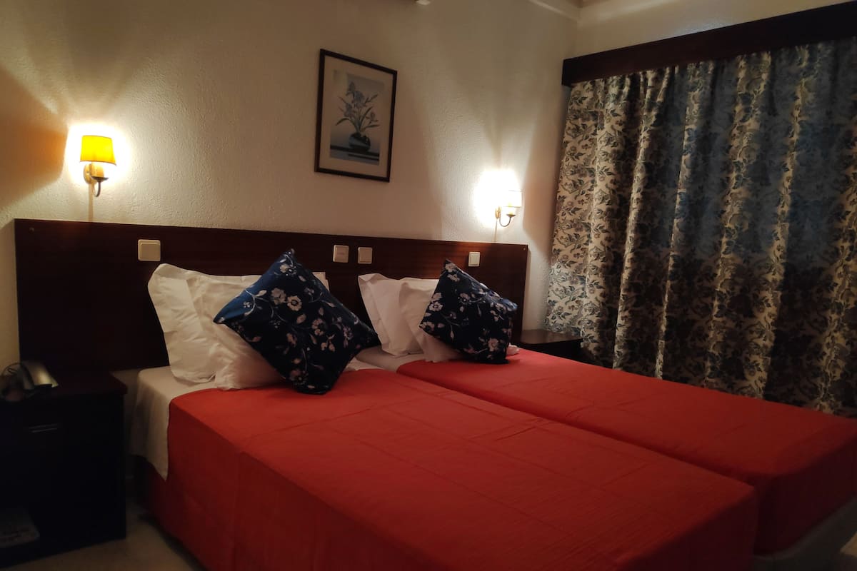 Une chambre d’hôtel avec un lit double, deux tables de chevet, une petite table, une lampe, un tableau encadré au mur et une fenêtre avec des rideaux.