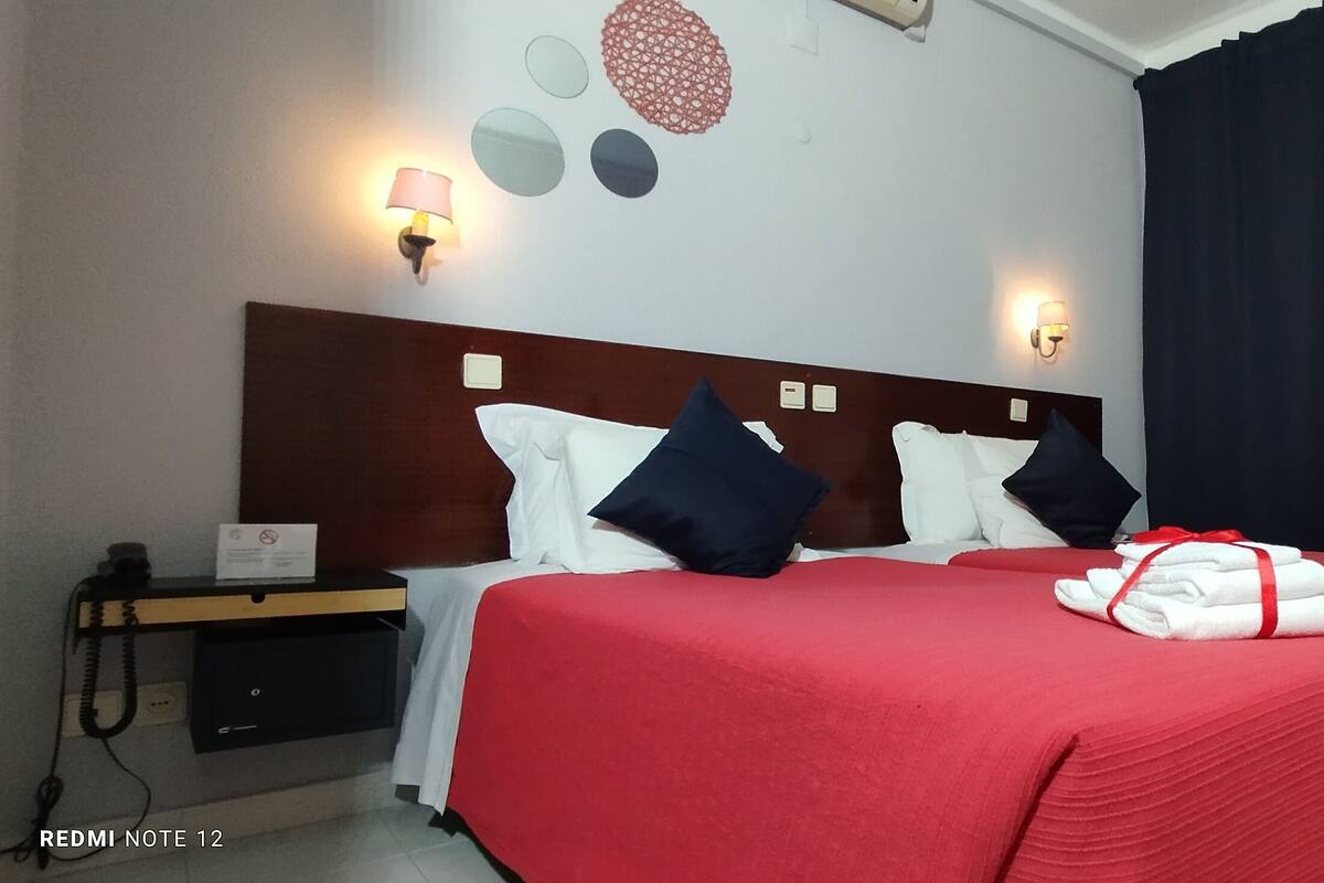 Une chambre d’hôtel avec deux lits, une literie rouge, une table de chevet avec un téléphone et des lampes fixées au mur.