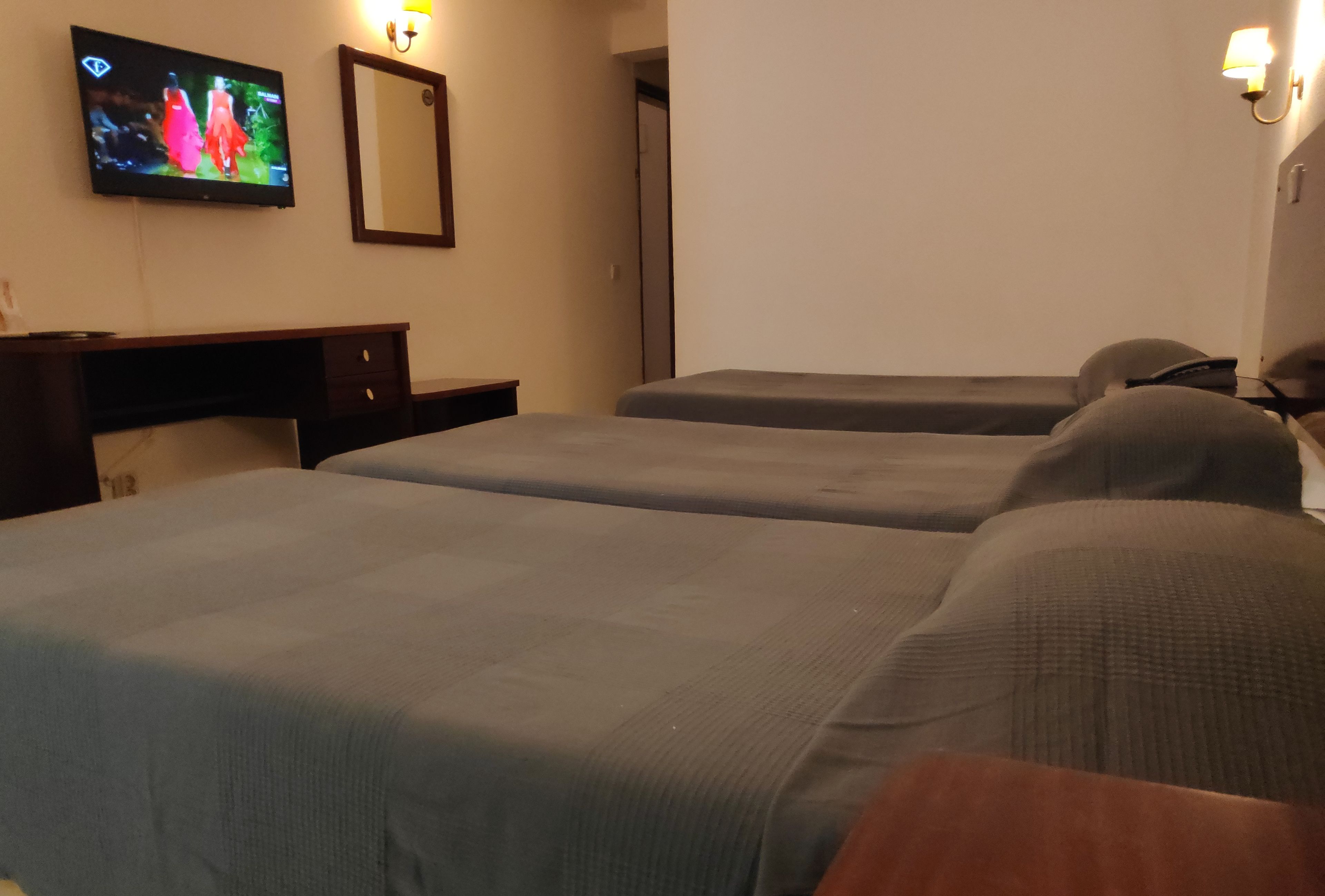 A hotel room with two single beds, a desk with a chair, a television mounted on the wall, and a mirror.