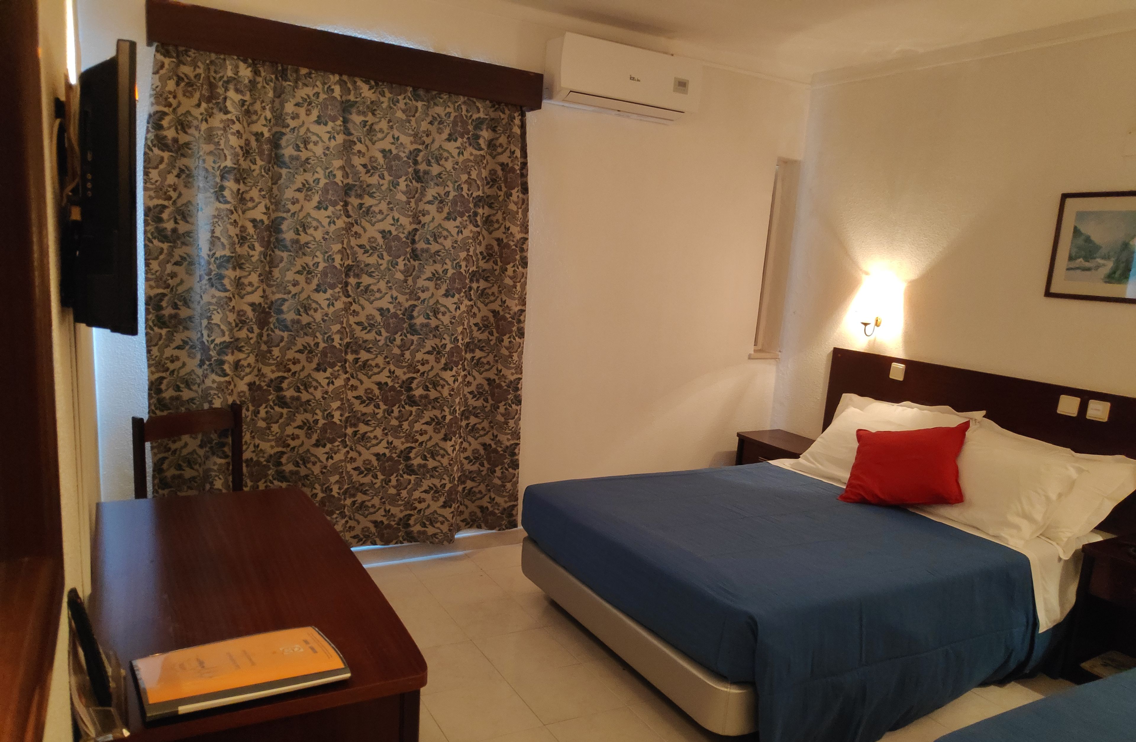 A hotel room with a bed, a desk, a chair, and an air conditioning unit.