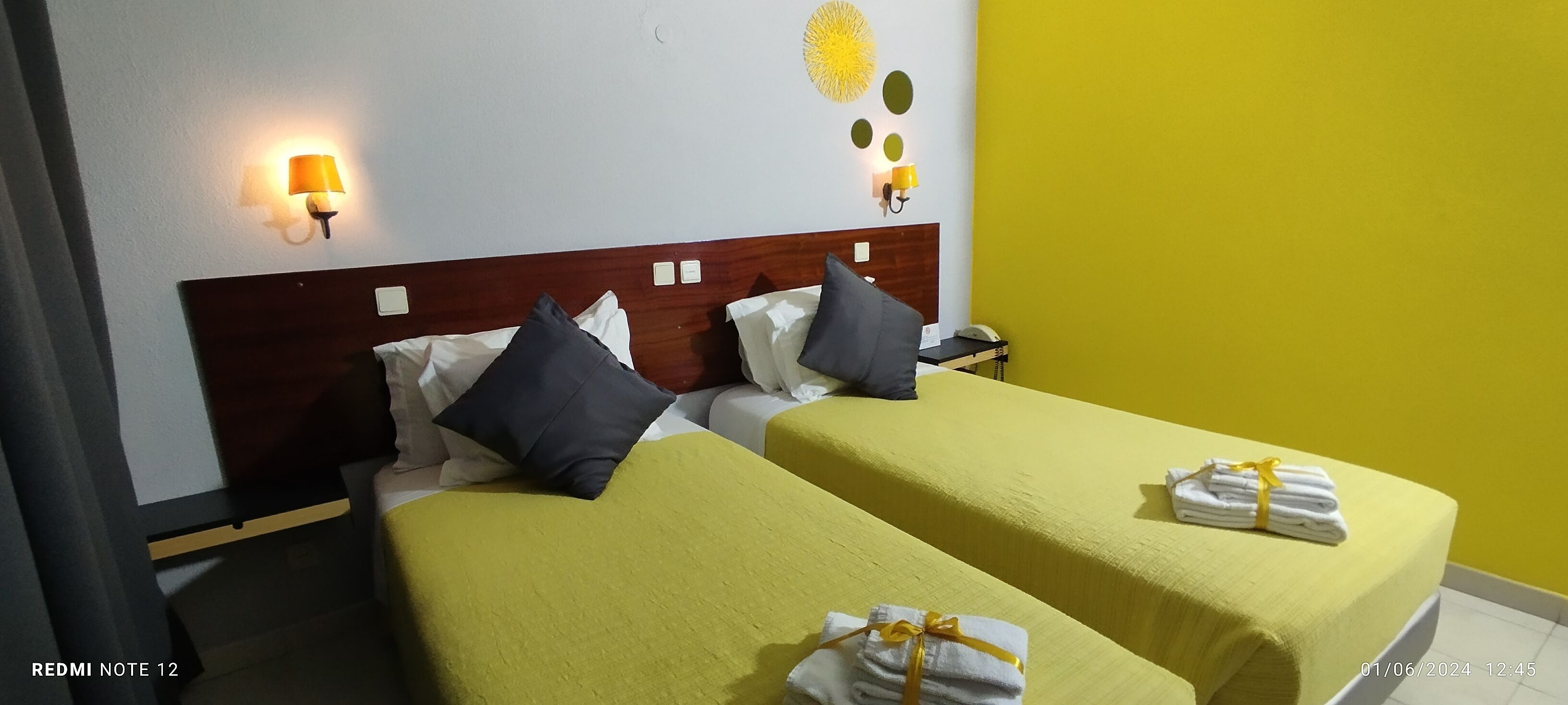 A hotel room with two beds, each with a yellow bedspread and pillows, a wooden headboard, bedside lamps, and a small table with a phone.