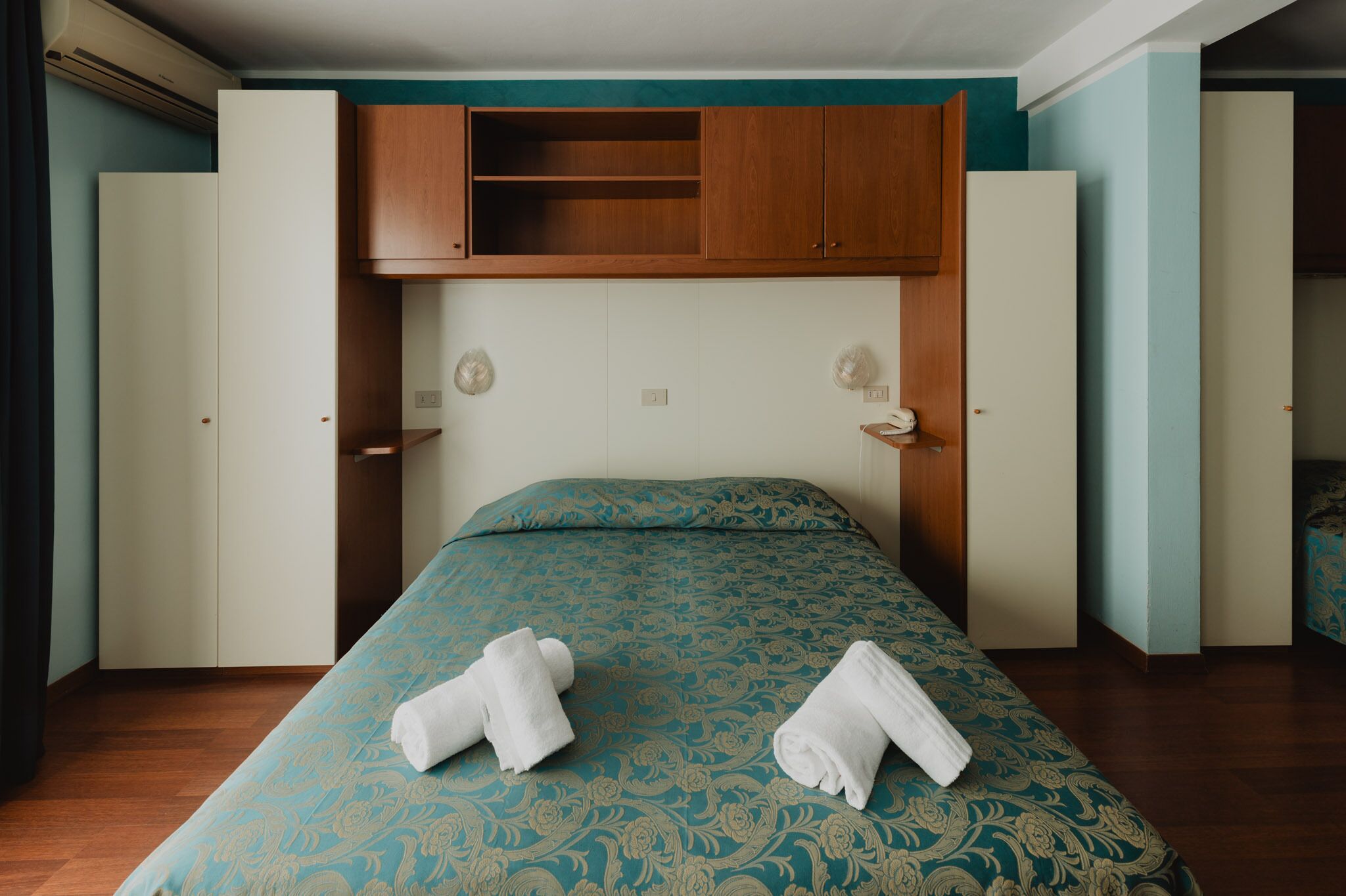 Habitación de hotel con una cama, dos puertas blancas, un armario de madera y una colcha de cama estampada en tonos verde agua.