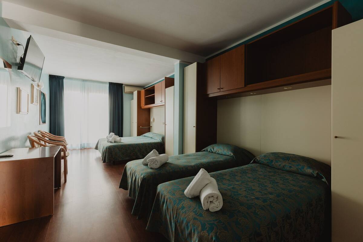 Una habitación de hotel con dos camas individuales, un escritorio de madera, una silla, un televisor montado en la pared y una ventana con cortinas.