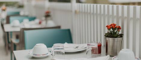 A dining table set with white plates, cups, and cutlery, a small potted plant with red flowers, and a metal bucket.