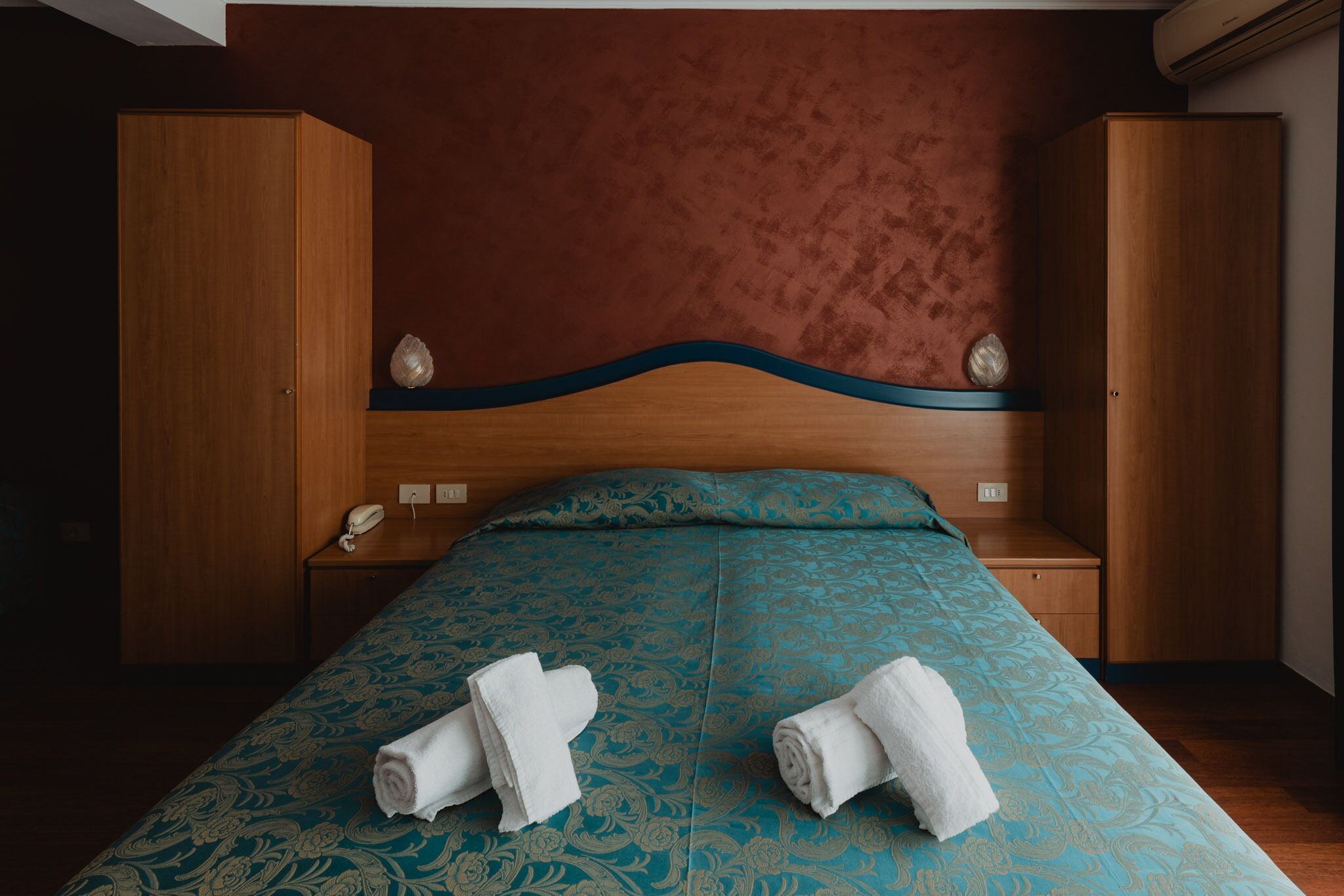 A hotel room with a bed, wooden wardrobe, bedside tables, and a telephone.