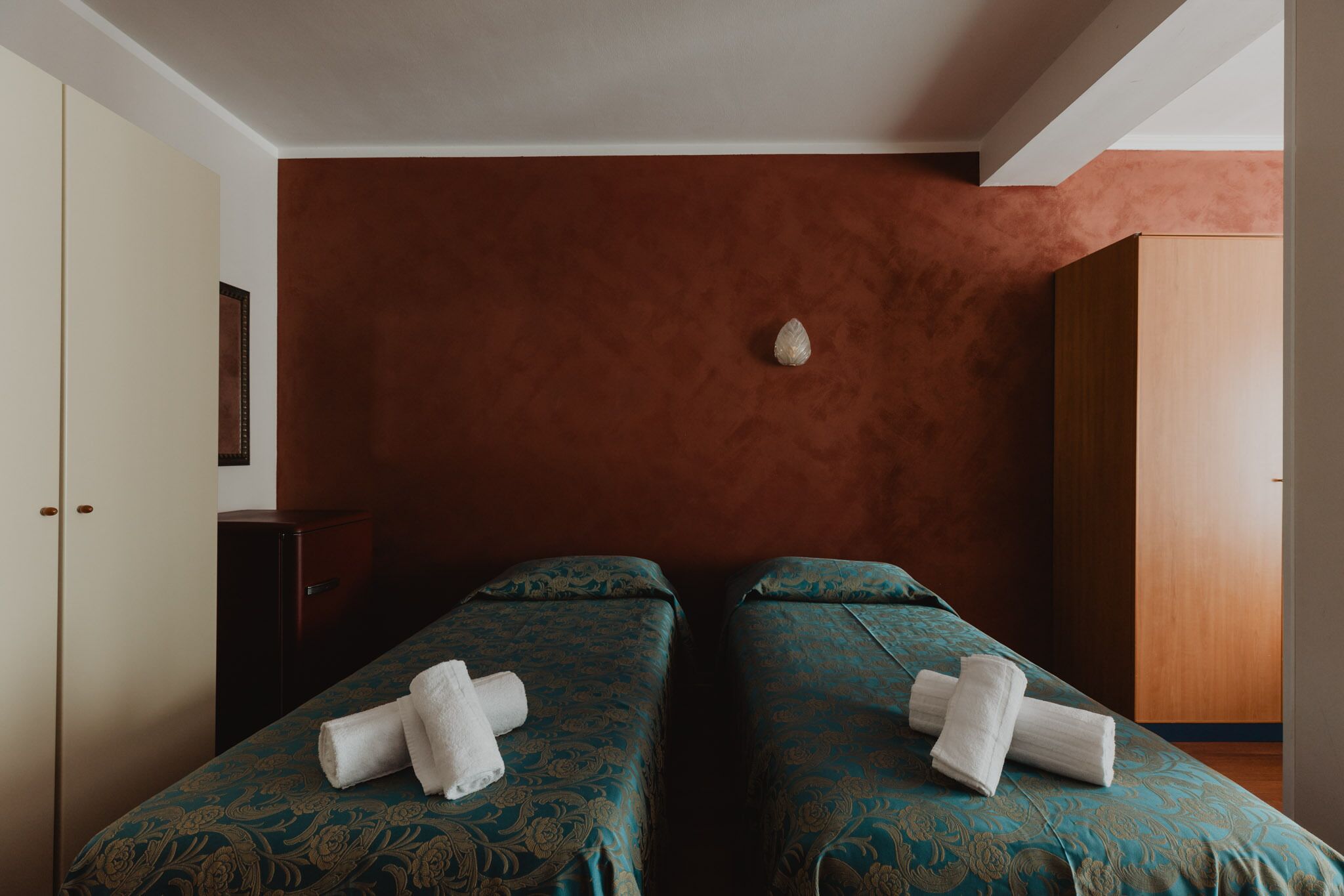 A room with two beds, each with a patterned bedspread and a rolled towel.