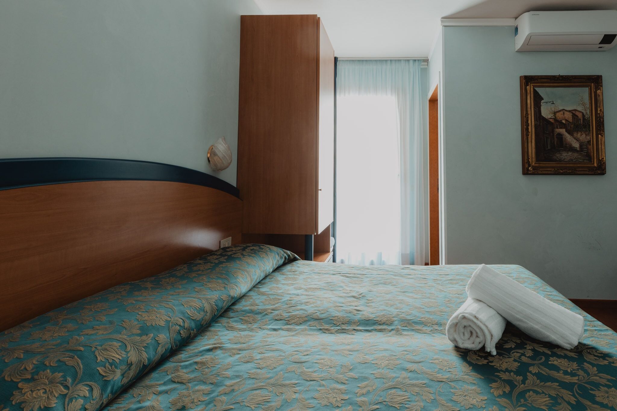 Ein Hotelzimmer mit einem hölzernen Kopfende, einer gemusterten Bettdecke, einem aufgerollten Handtuch, einem Gemälde an der Wand und einem Fenster mit Vorhängen.