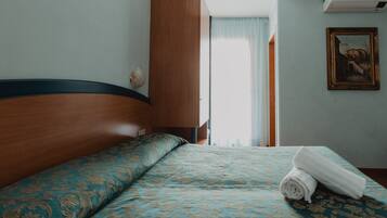 Ein Hotelzimmer mit einem hölzernen Kopfende, einer gemusterten Bettdecke, einem aufgerollten Handtuch, einem Gemälde an der Wand und einem Fenster mit Vorhängen.