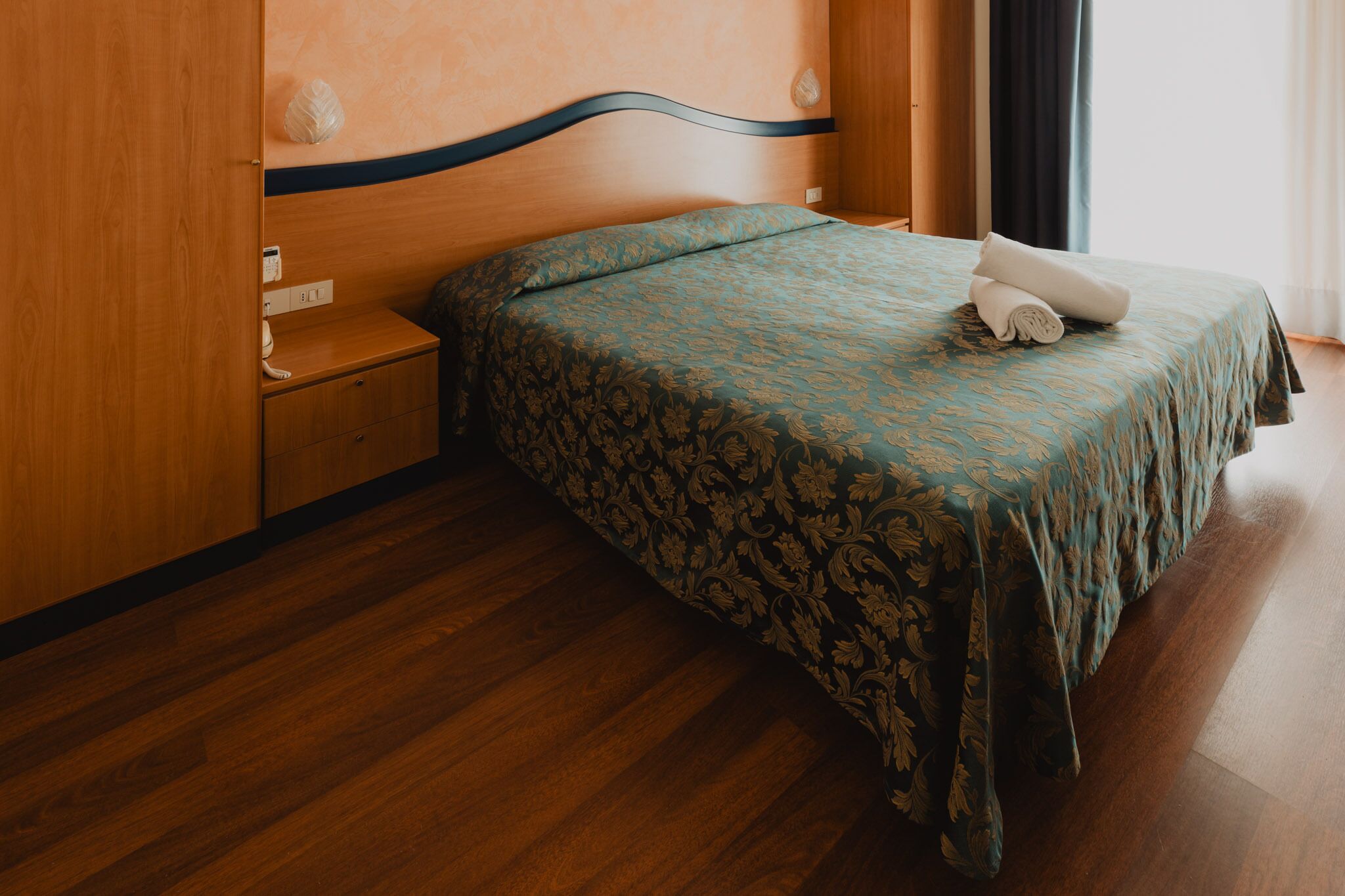 Una cama bien tendida con una colcha estampada y una toalla enrollada, una mesita de noche de madera con una lámpara y un armario de madera.
