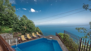 A rectangular swimming pool with a wooden deck, surrounded by a metal fence and trees. - Nido degli Dei (Agerola)