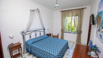 Un dormitorio con cama, mesita de noche, silla, escritorio, televisor y ventana con cortinas.