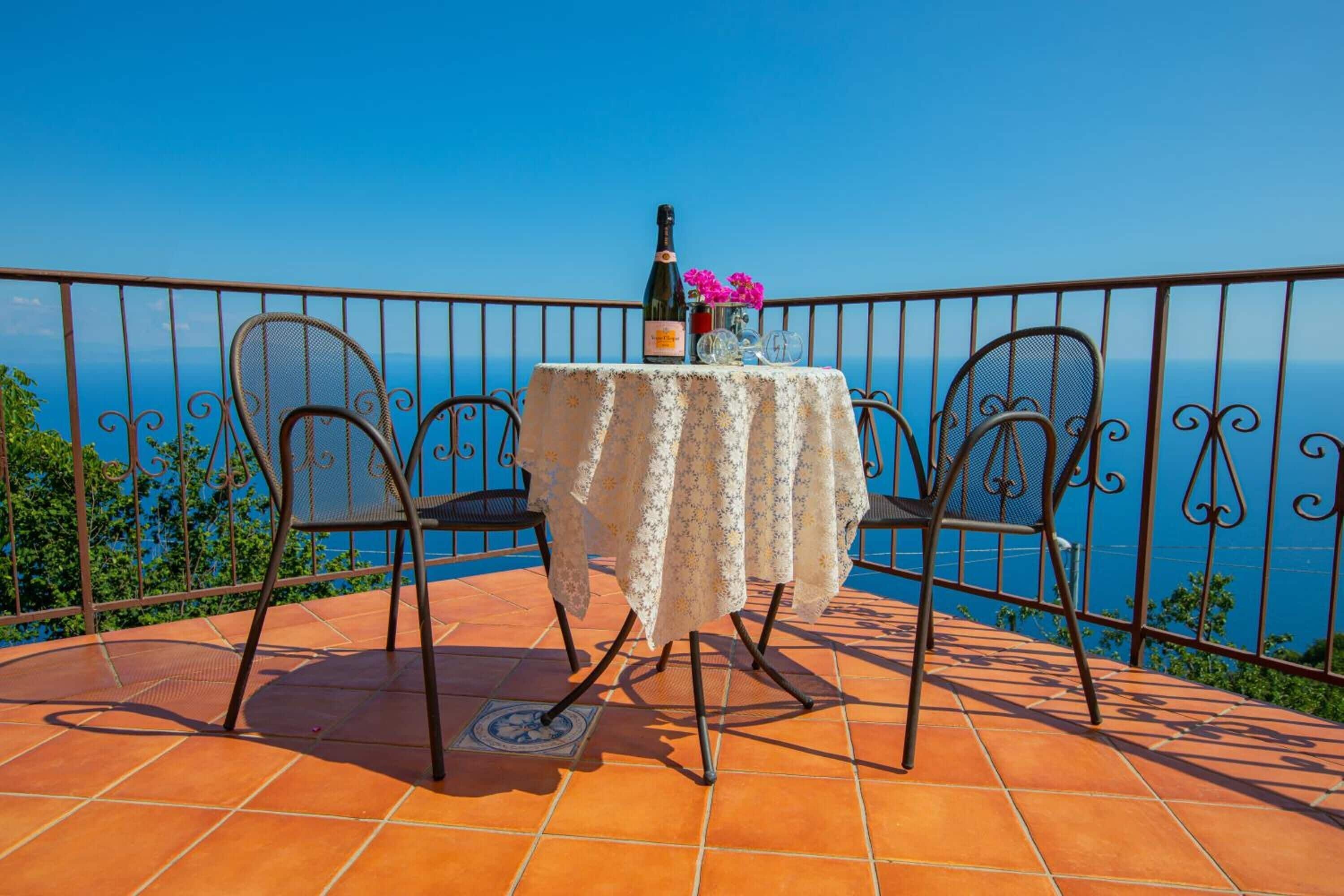 A terrace with a table set for two, a bottle of wine, and pink flowers.