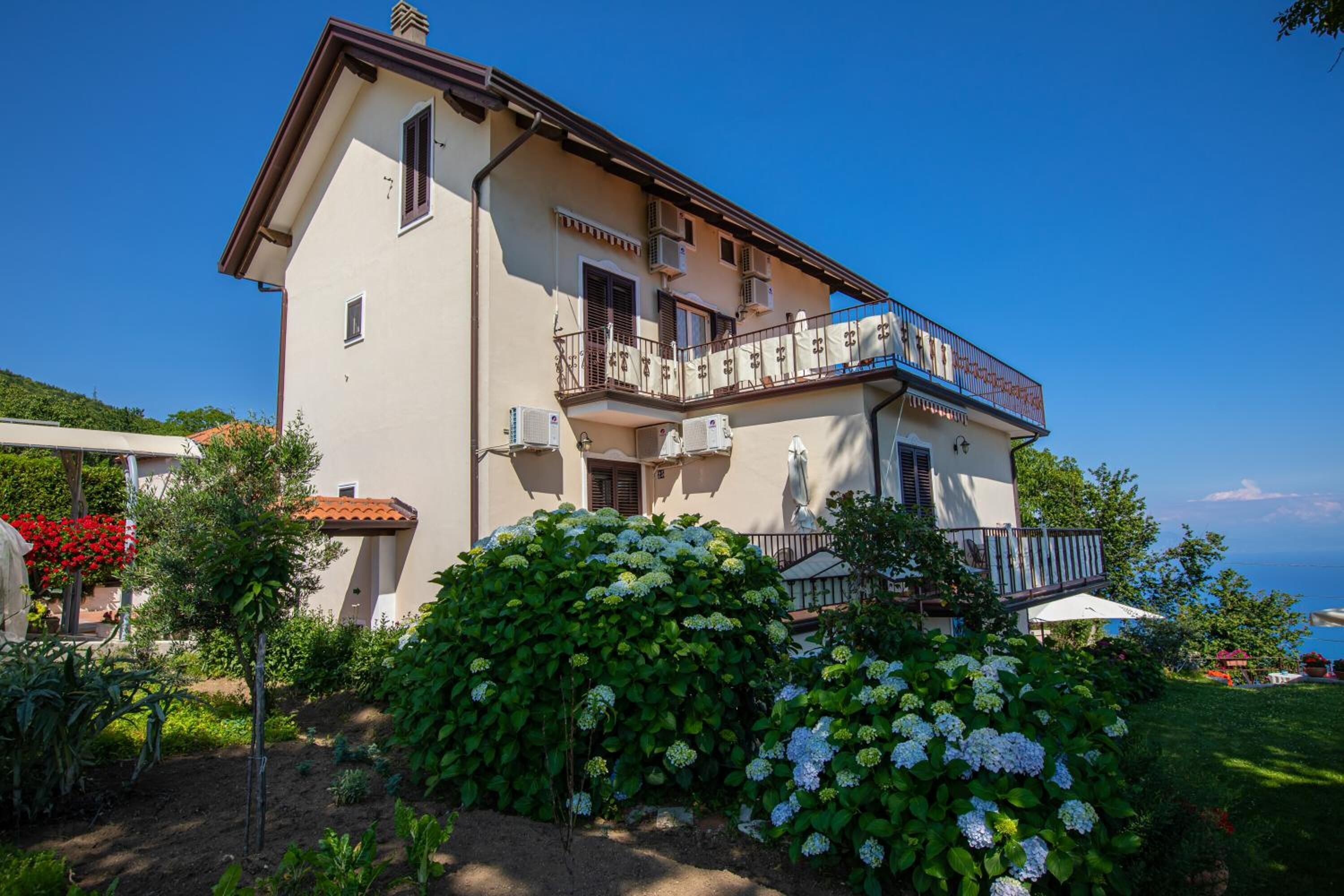 Un immeuble de deux étages doté d’un balcon, de climatiseurs et d’un jardin où poussent des hortensias.