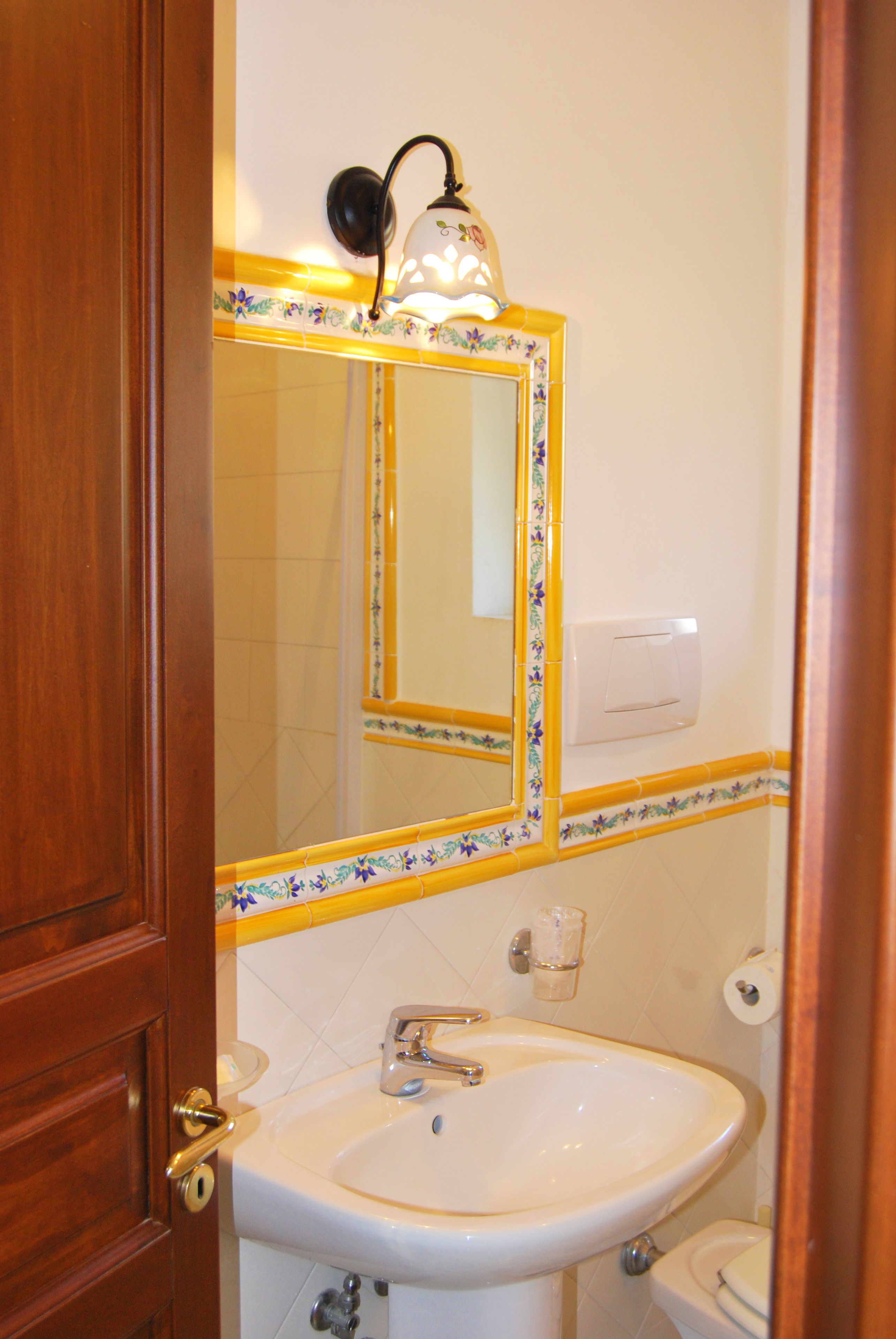 A bathroom with a wooden door, a white sink, a mirror with a floral border, a light fixture, and a toilet.
